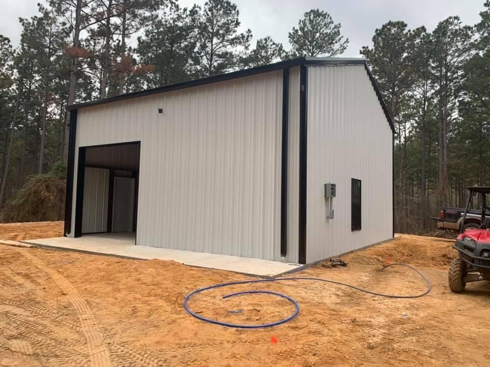 A white metal building with a large open door, black trim, and two windows in a wooded setting.