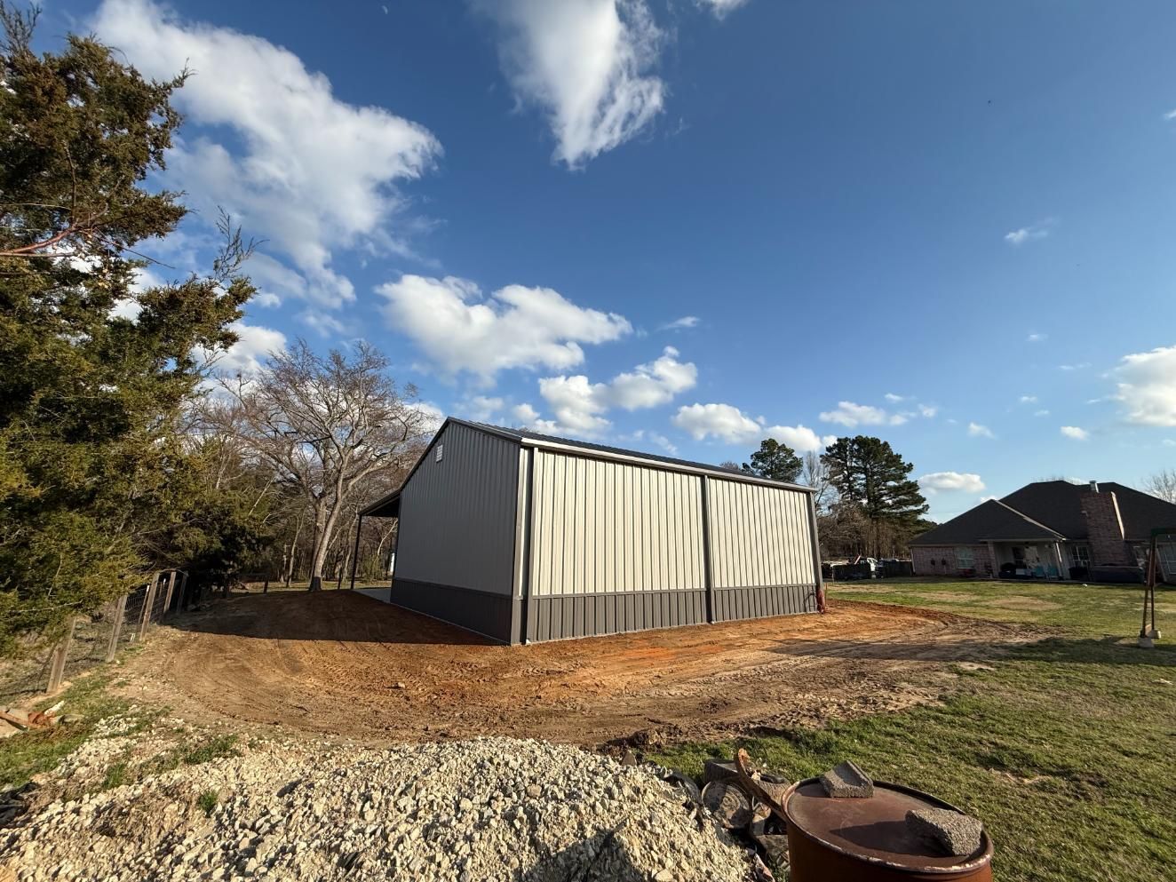 Metal shed under a blue sky with scattered clouds, set in a grassy area with a dirt road.