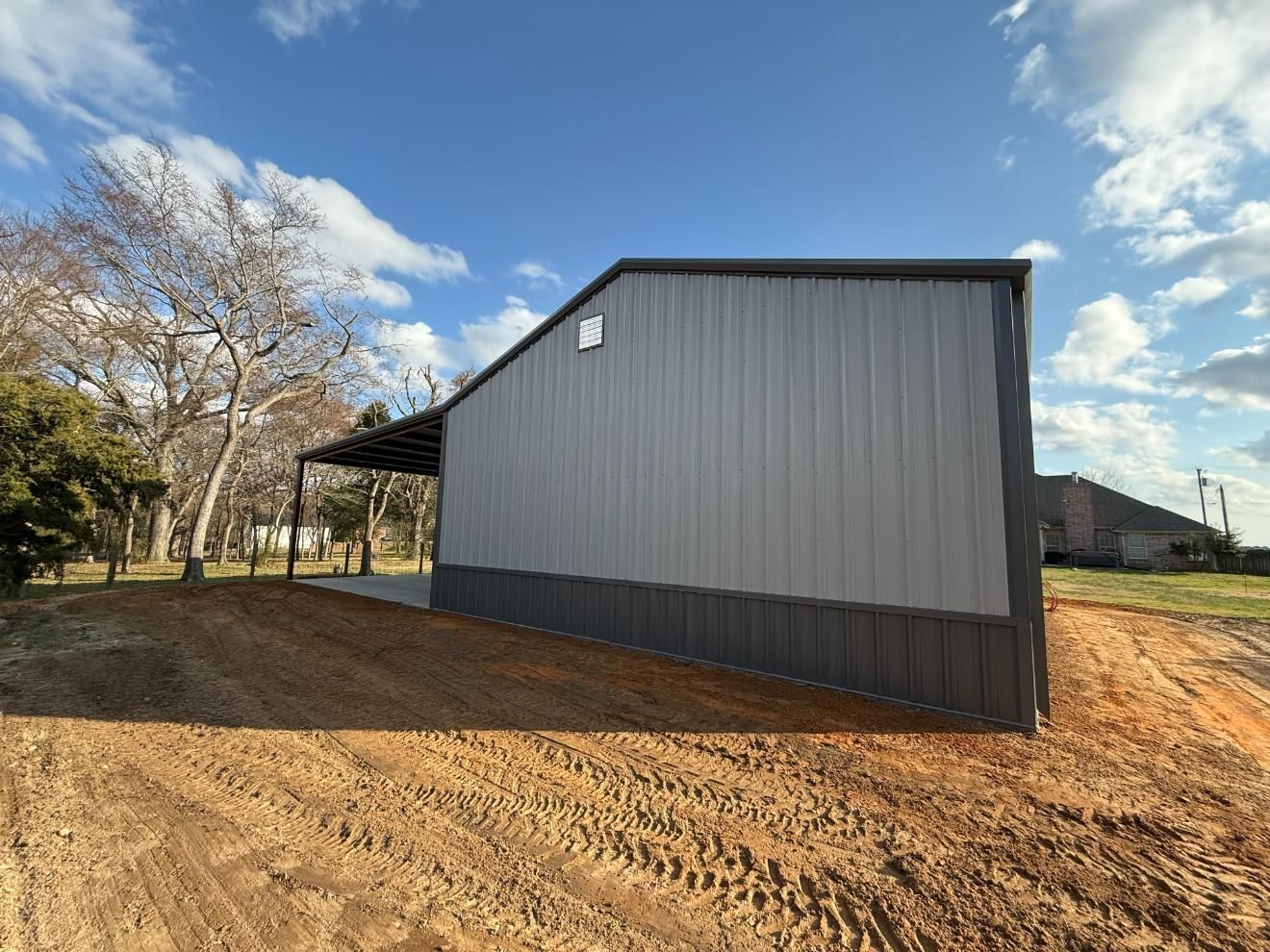 A gray metal barn with a dark bottom panel, set on a brown dirt lot under a blue sky.
