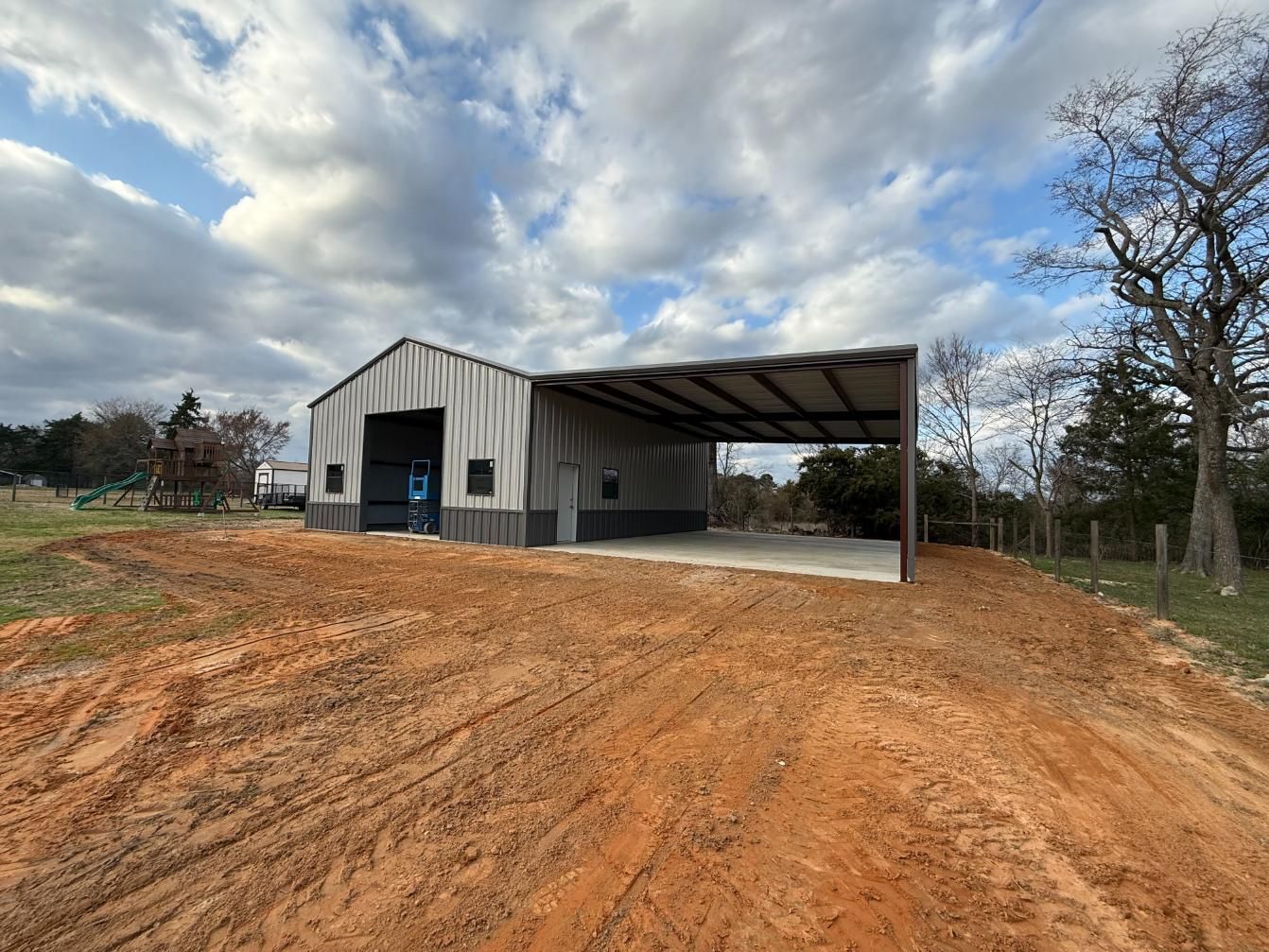 Metal barn with open carport, on a dirt lot under cloudy skies.