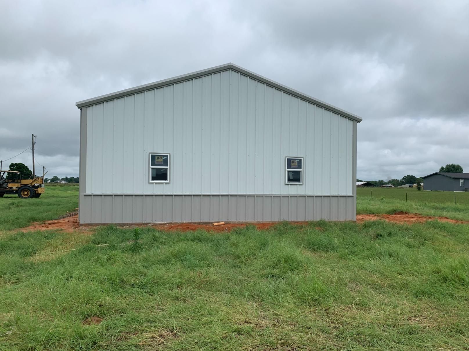 White and gray metal building with two windows, set in a grassy field under a cloudy sky.