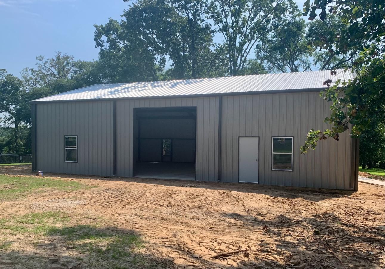 Gray metal storage building with large door, small windows, and a white door, on a dirt lot.
