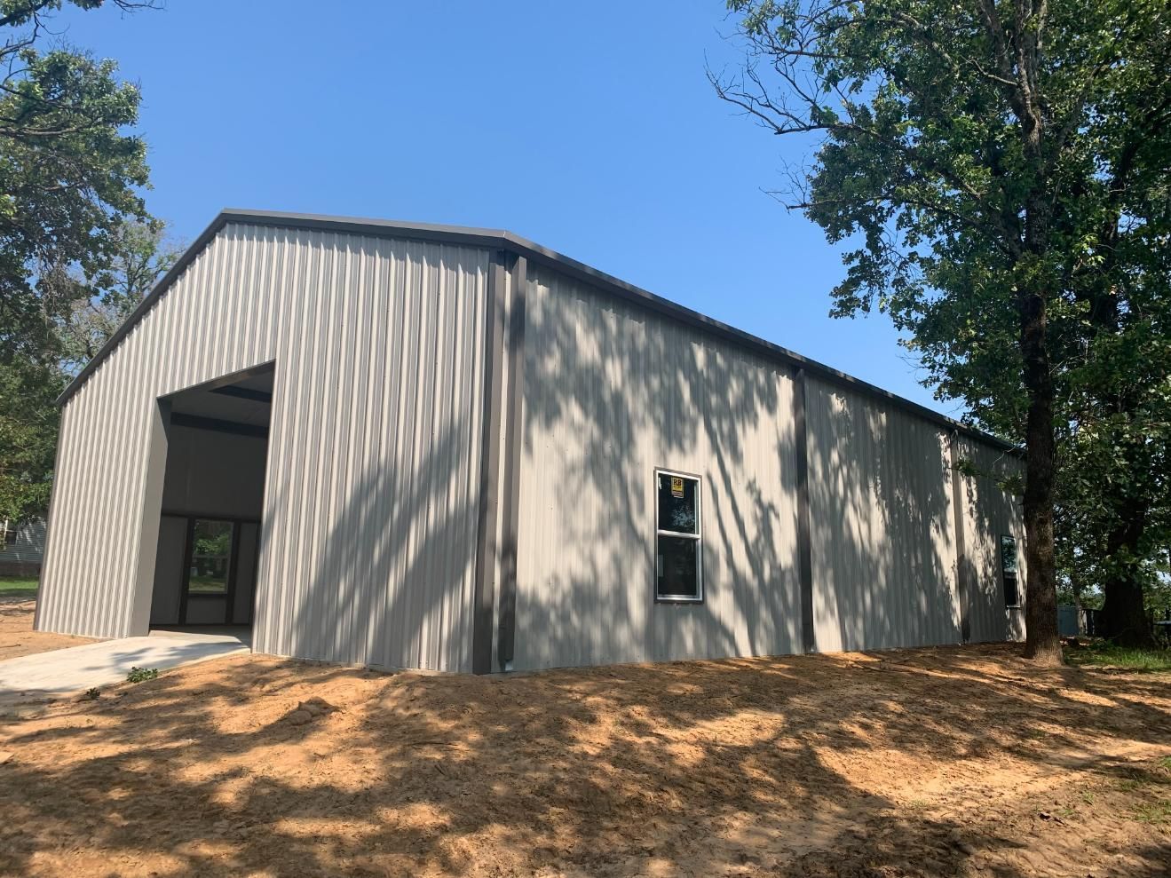 Metal building with open bay door, window, and surrounding trees under a blue sky.