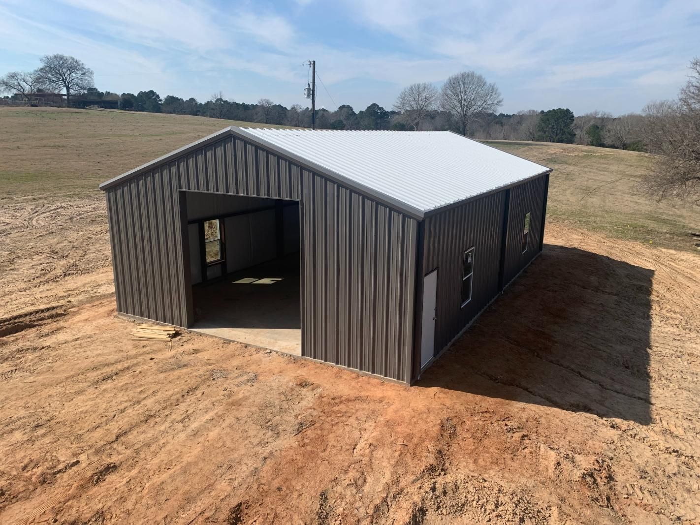 Metal building with open bay and door on a dirt lot, gray walls, white roof, and blue sky.