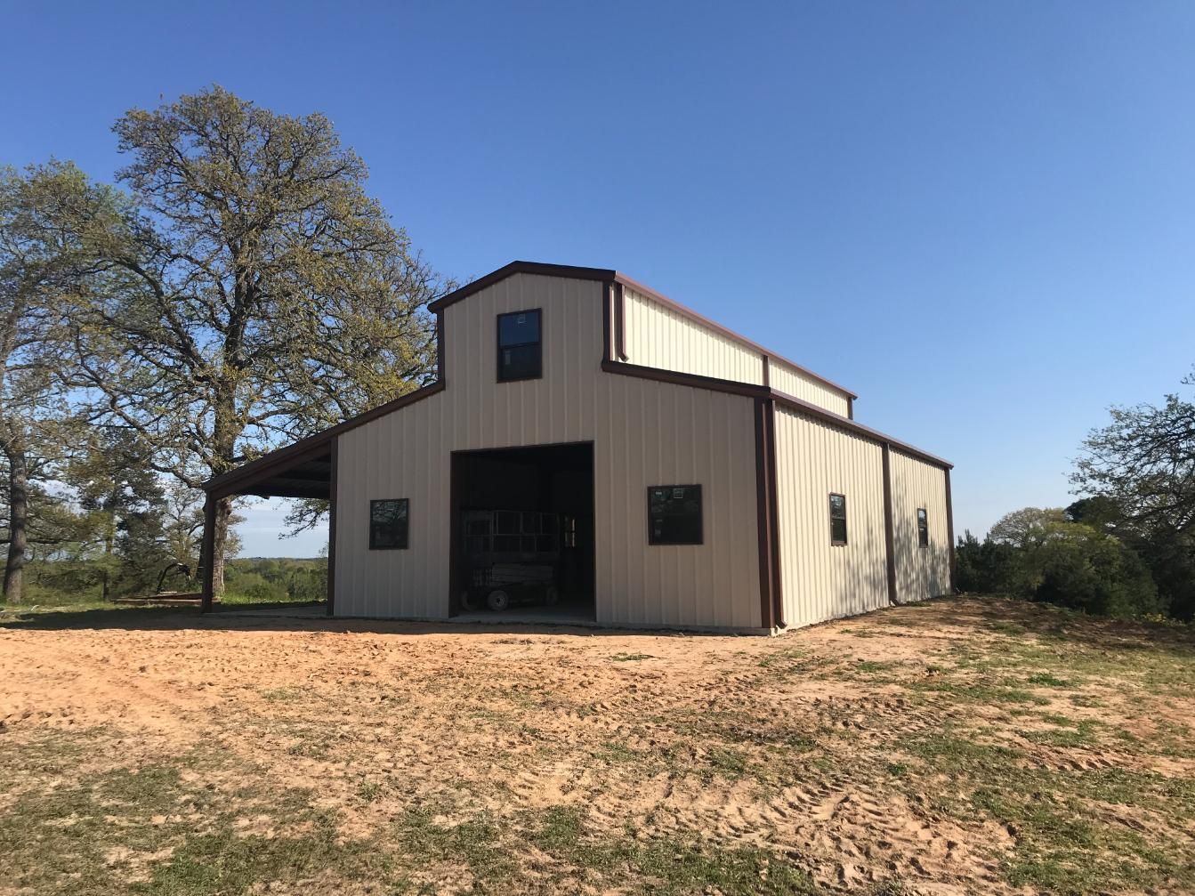 Tan metal barn with brown trim, windows, and awning, set against a blue sky and dry, brown field.