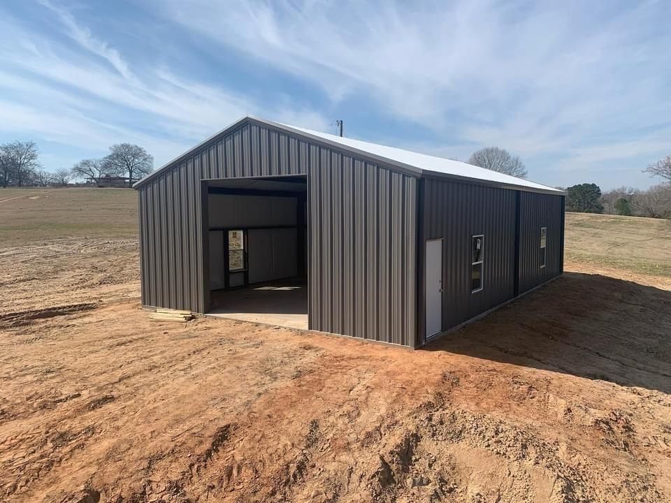 Metal barn with dark brown siding, white roof, and open garage door on a dirt lot under a blue sky.