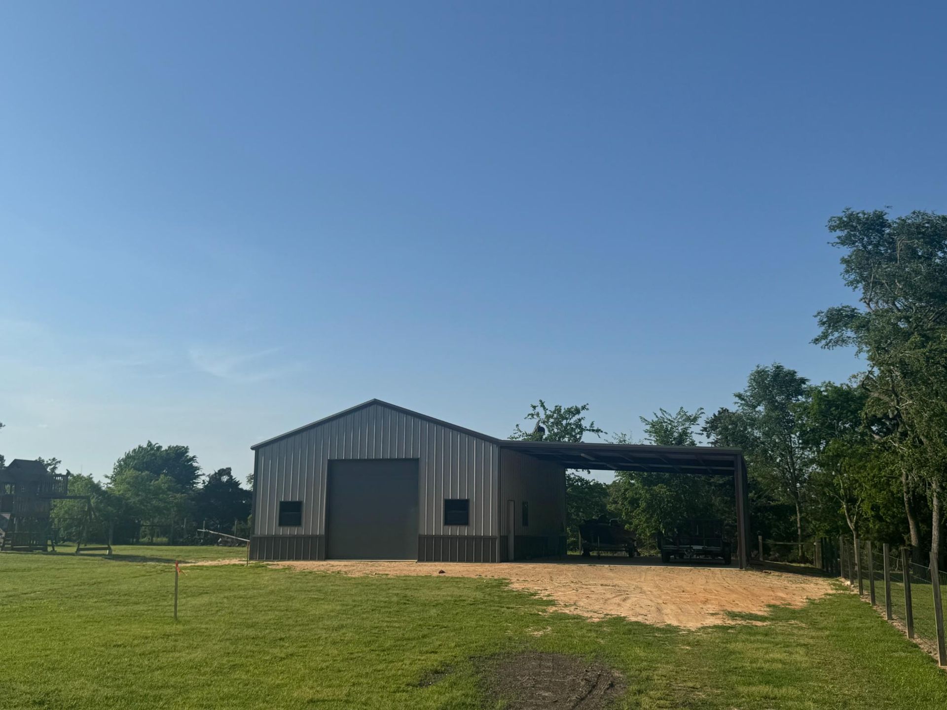 Metal barn with a covered side area, on green grass, against a blue sky.
