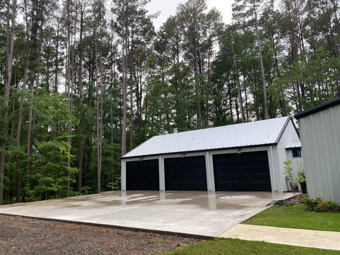 A gray, concrete carport with three bays, a white roof, and a forest background.