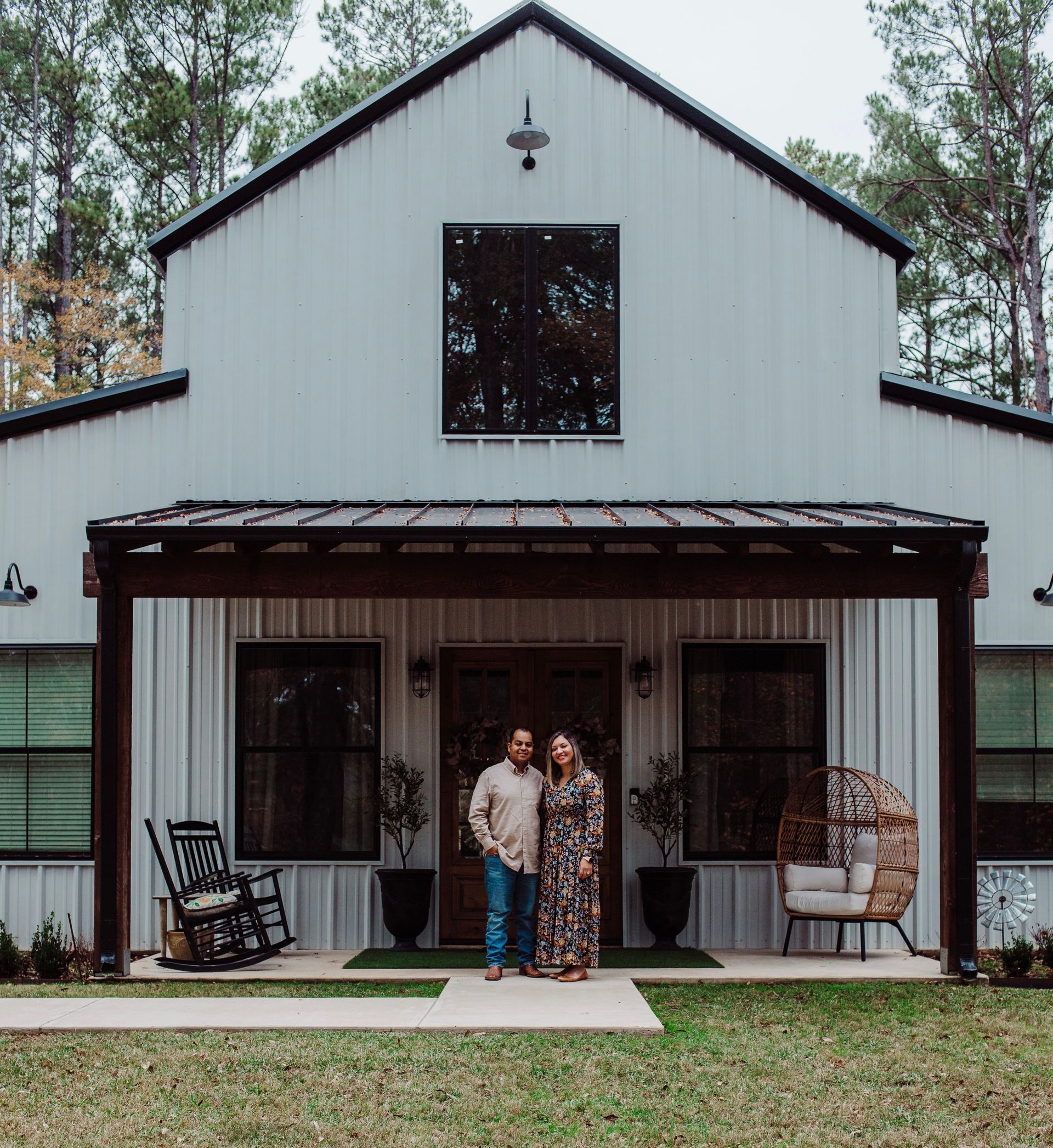 Couple standing in front of a white barn with a porch, rocking chair, and chair.
