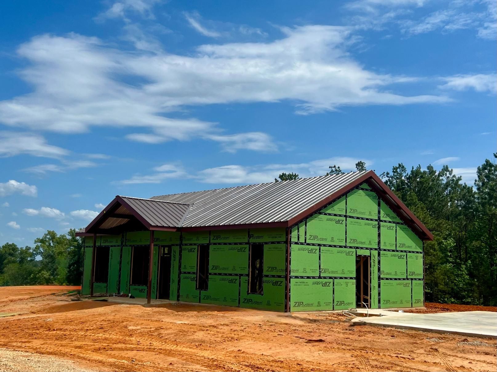 House under construction with brown metal roof, green wrap, and brown trim. Sunny day, blue sky.