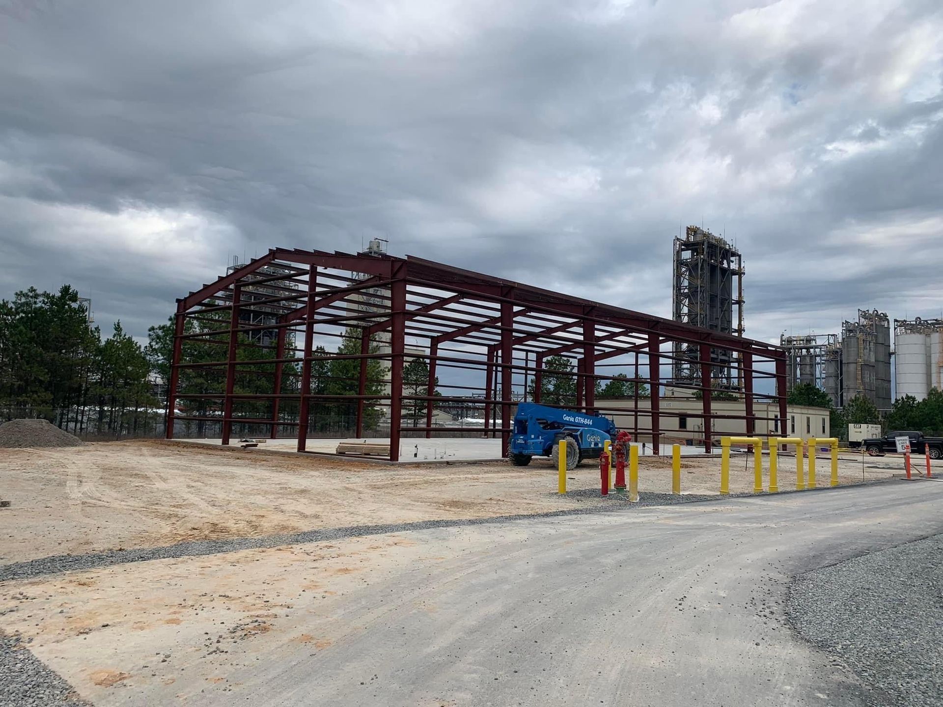 Steel frame of a building under construction; blue lift; dirt ground and cloudy sky.