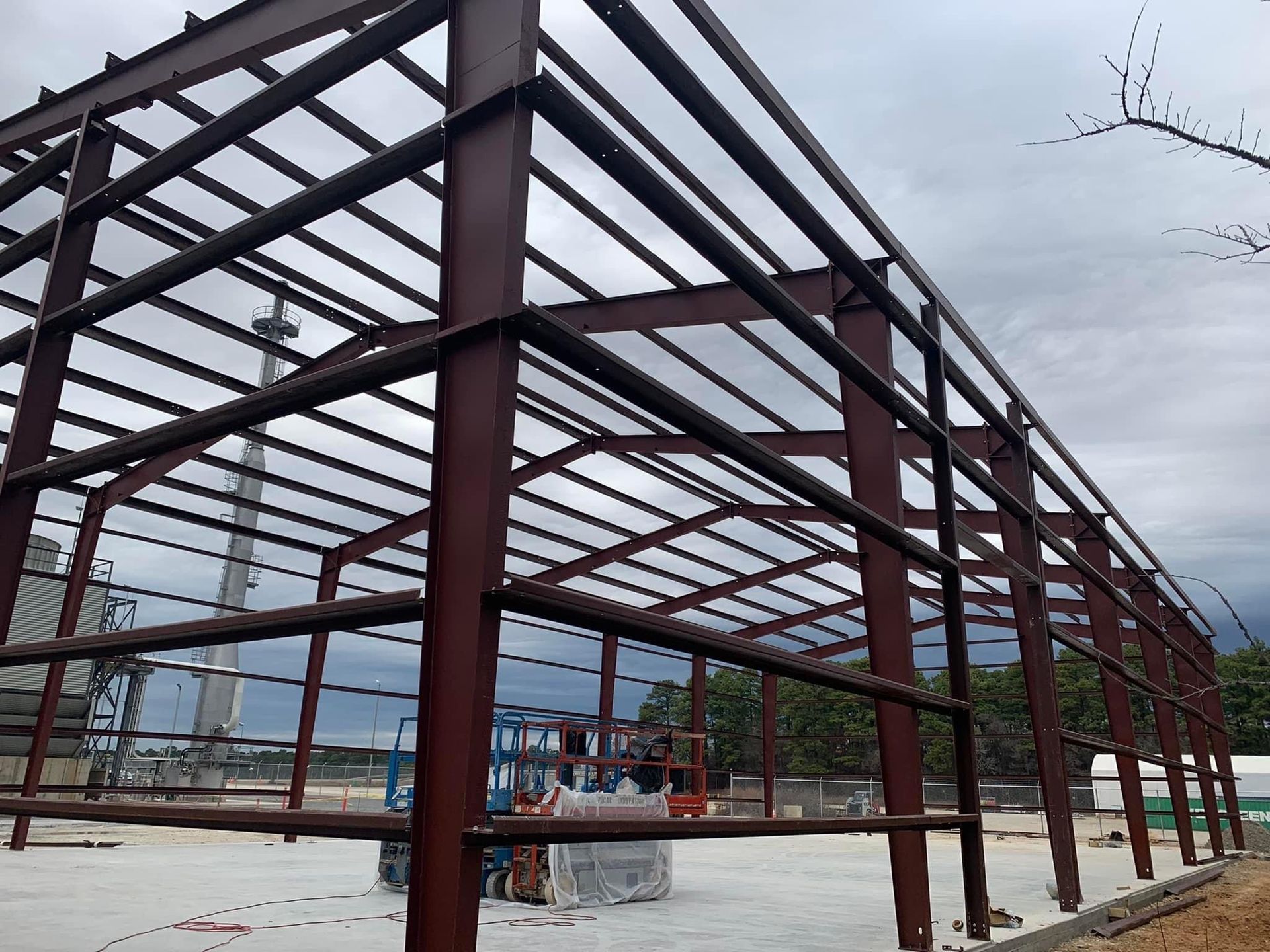 Steel frame of a building under construction; brown beams against a cloudy sky; a lift and concrete base are visible.