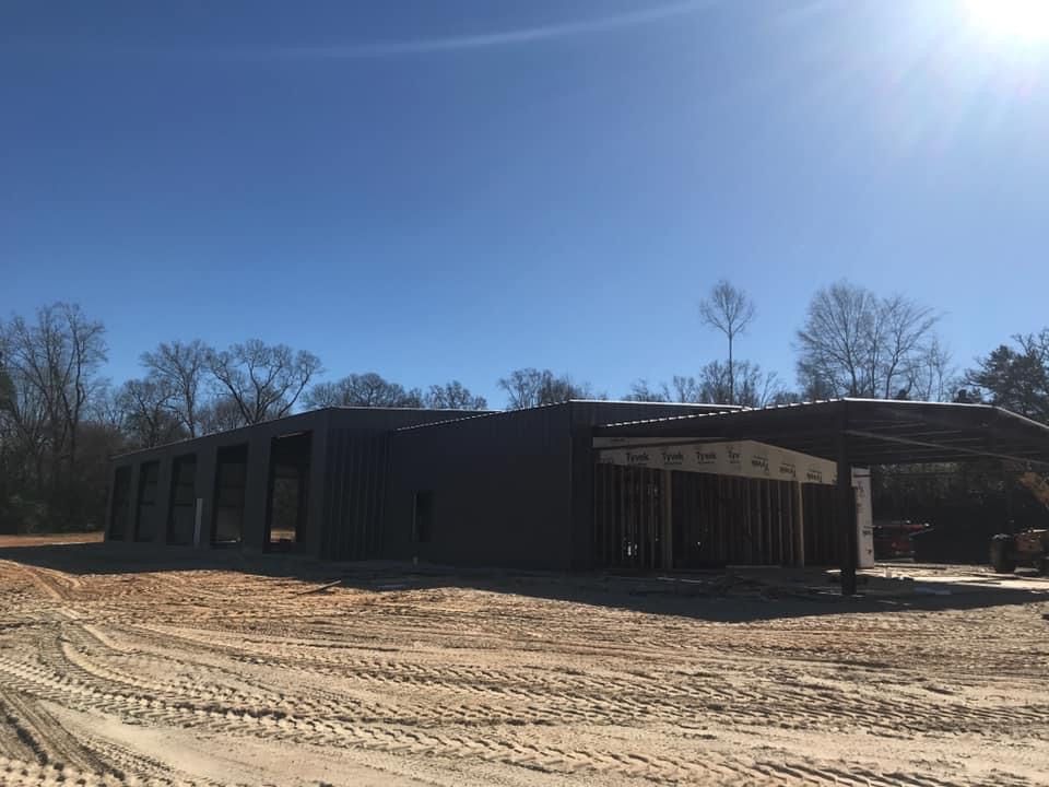 Construction site; large dark gray metal building under a blue sky, surrounded by dirt and trees.