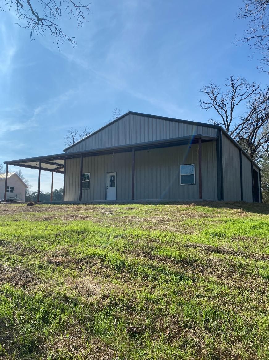 Metal building with a porch, situated on a grassy hill. Blue sky with trees in the background.