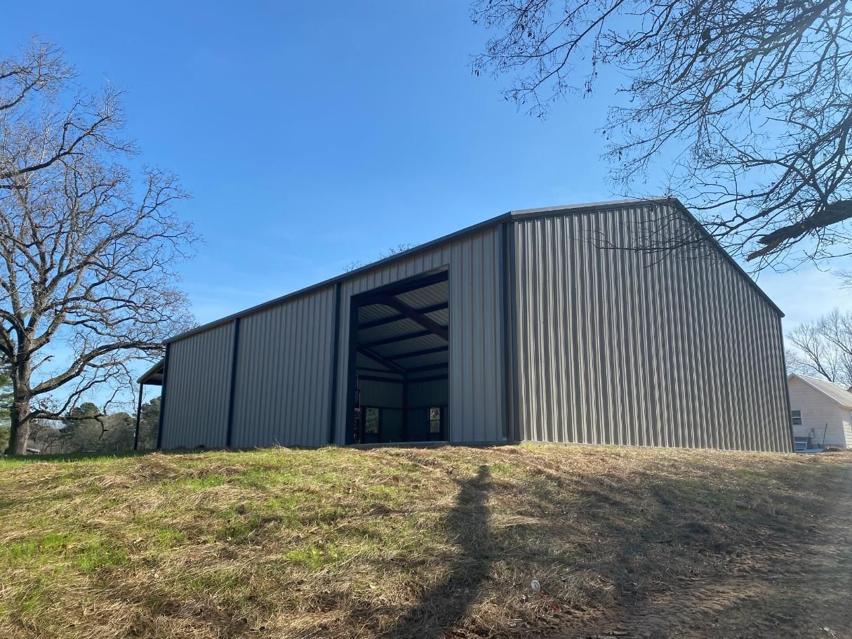 Large gray metal barn on a grassy hill under a blue sky, trees frame the view.
