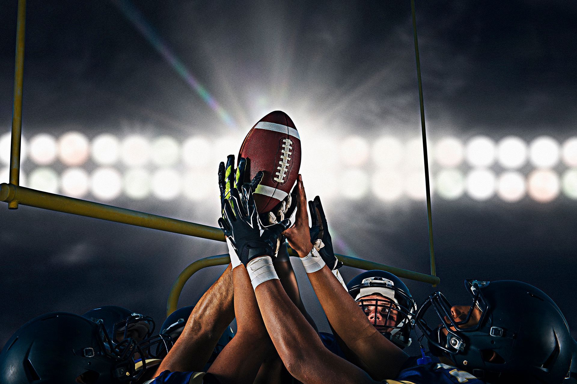 Football players reaching up to catch a ball under bright stadium lights