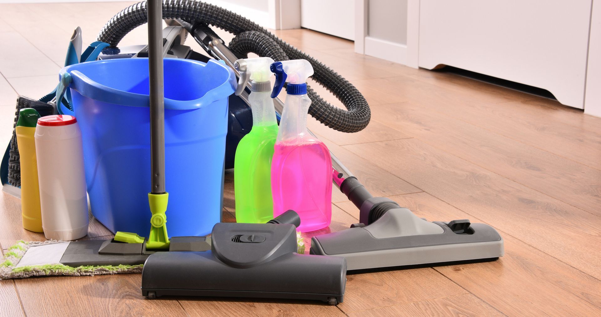 Cleaning supplies on wood floor, including a bucket, spray bottles, vacuum, and mop.