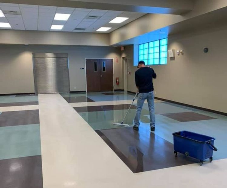 A person mops a large room with patterned flooring. The room has neutral-colored walls, large doors, and a window.