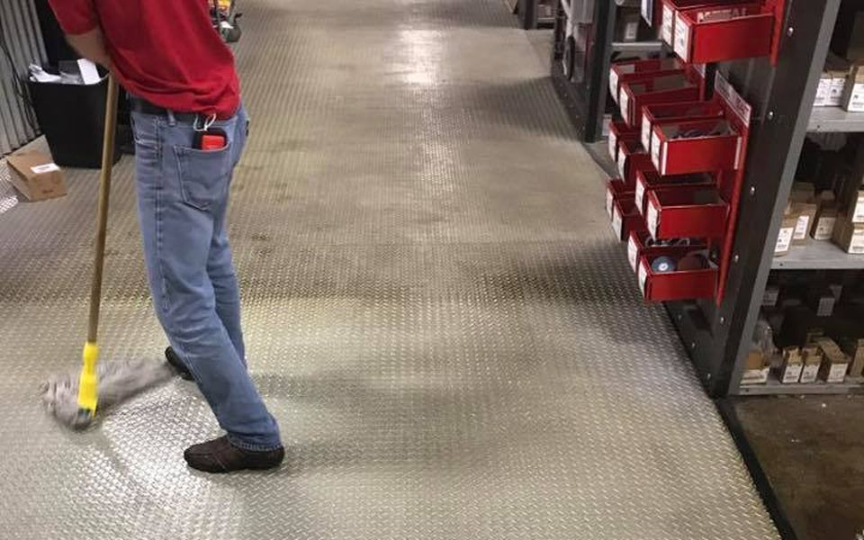 A person in jeans and a red shirt sweeps a warehouse floor.