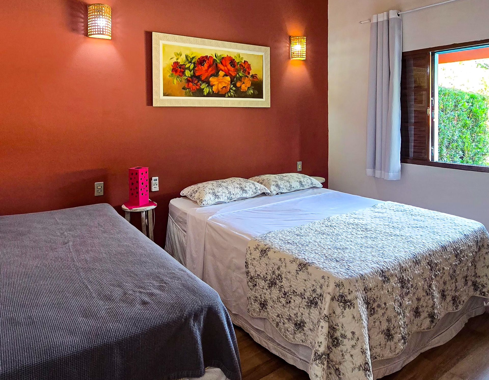 Quarto com duas camas encostadas em uma parede vermelha. Quadro floral acima da cama de casal, janela com vista para o verde.