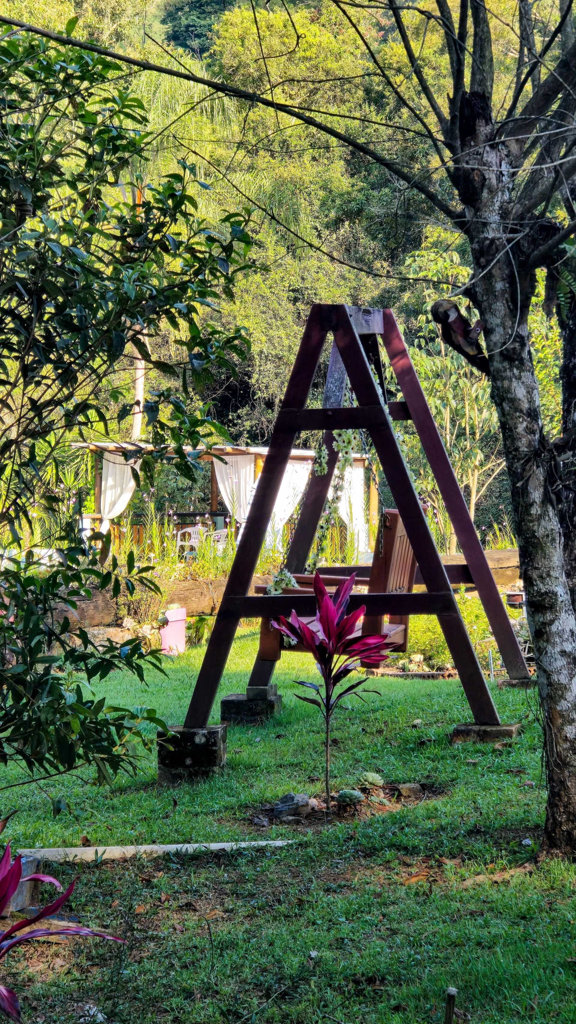 Um balanço de madeira em um gramado, com árvores e um gazebo coberto ao fundo.