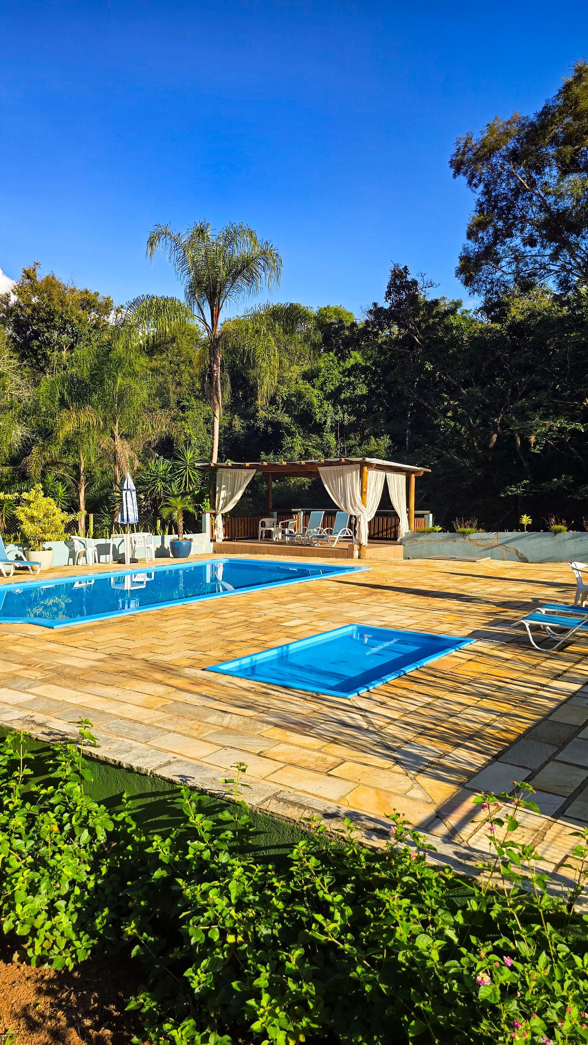 Duas piscinas azuis com um gazebo, rodeadas por árvores sob um céu azul brilhante.