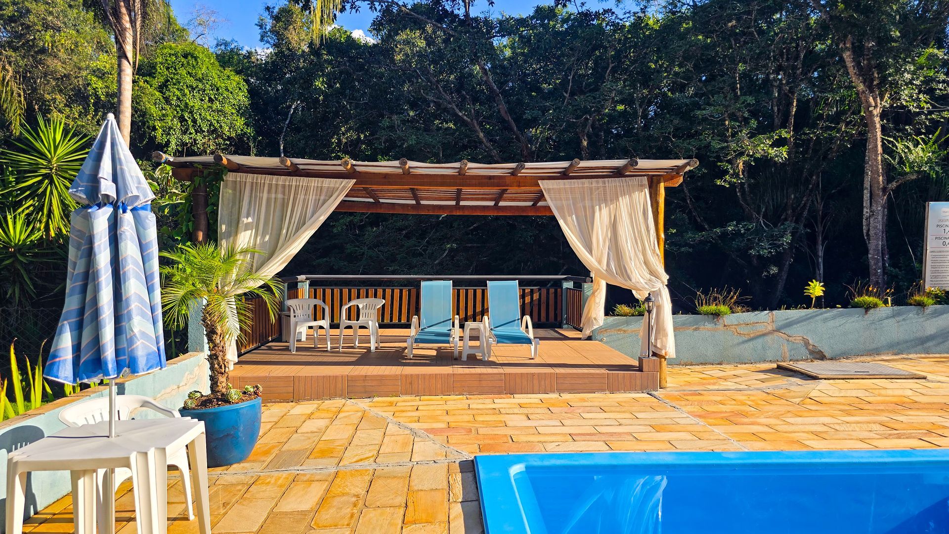 Gazebo à beira da piscina com cortinas drapeadas e espreguiçadeiras azuis. Guarda-sol azul e mesa em primeiro plano.