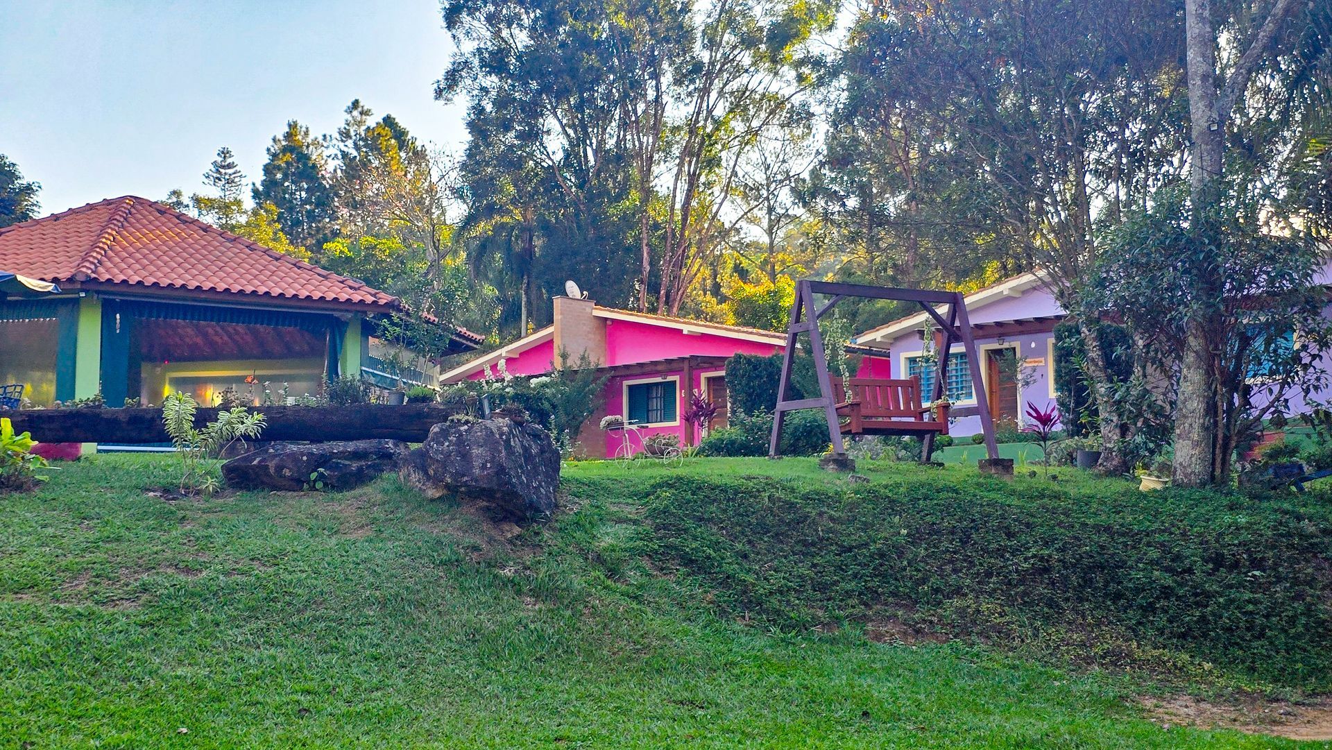 Casas com fachadas coloridas, telhados vermelhos e rosa, em um gramado verde com árvores ao fundo.