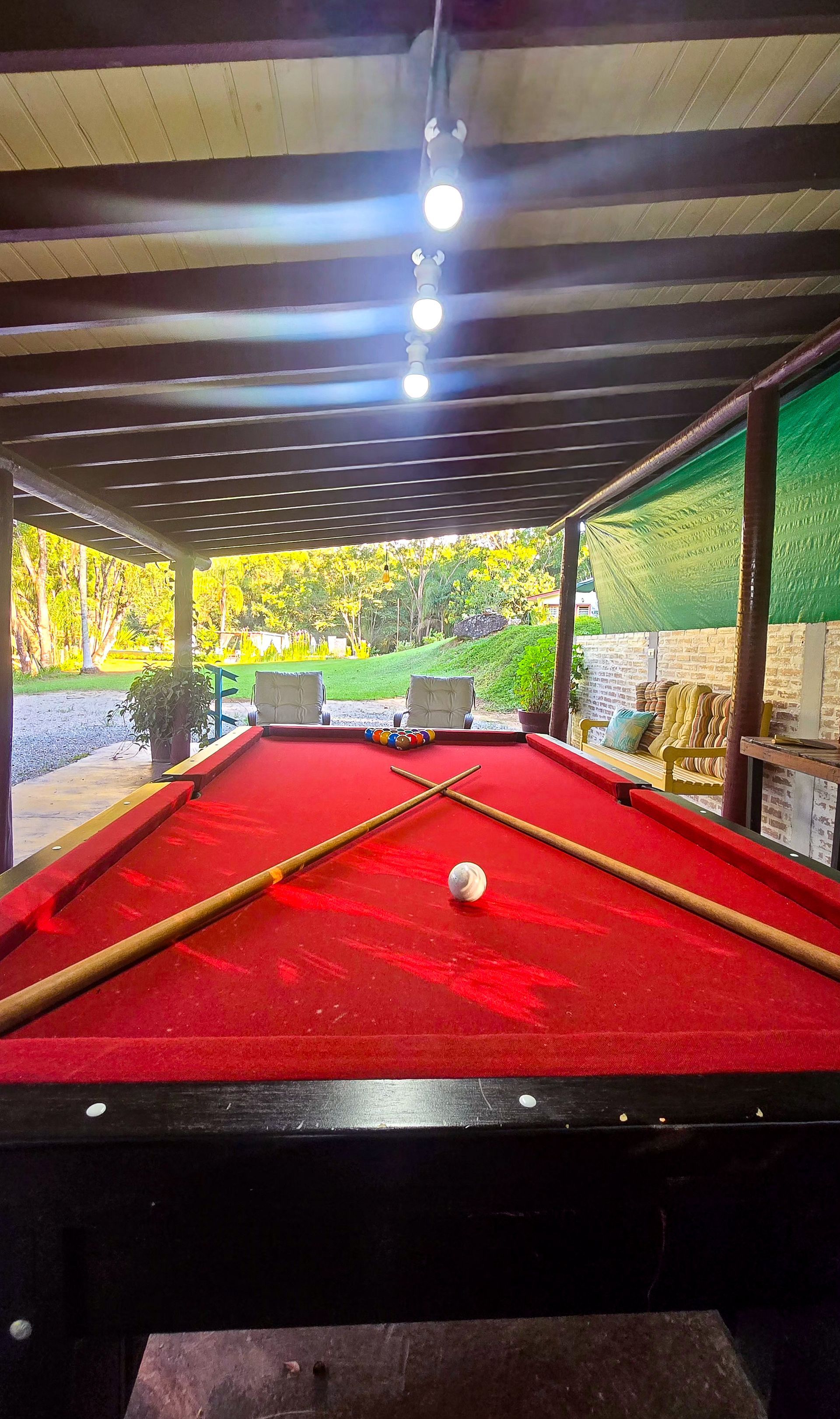 Mesa de bilhar com feltro vermelho sob uma pérgola de madeira, tacos cruzados e uma bola branca ao centro.