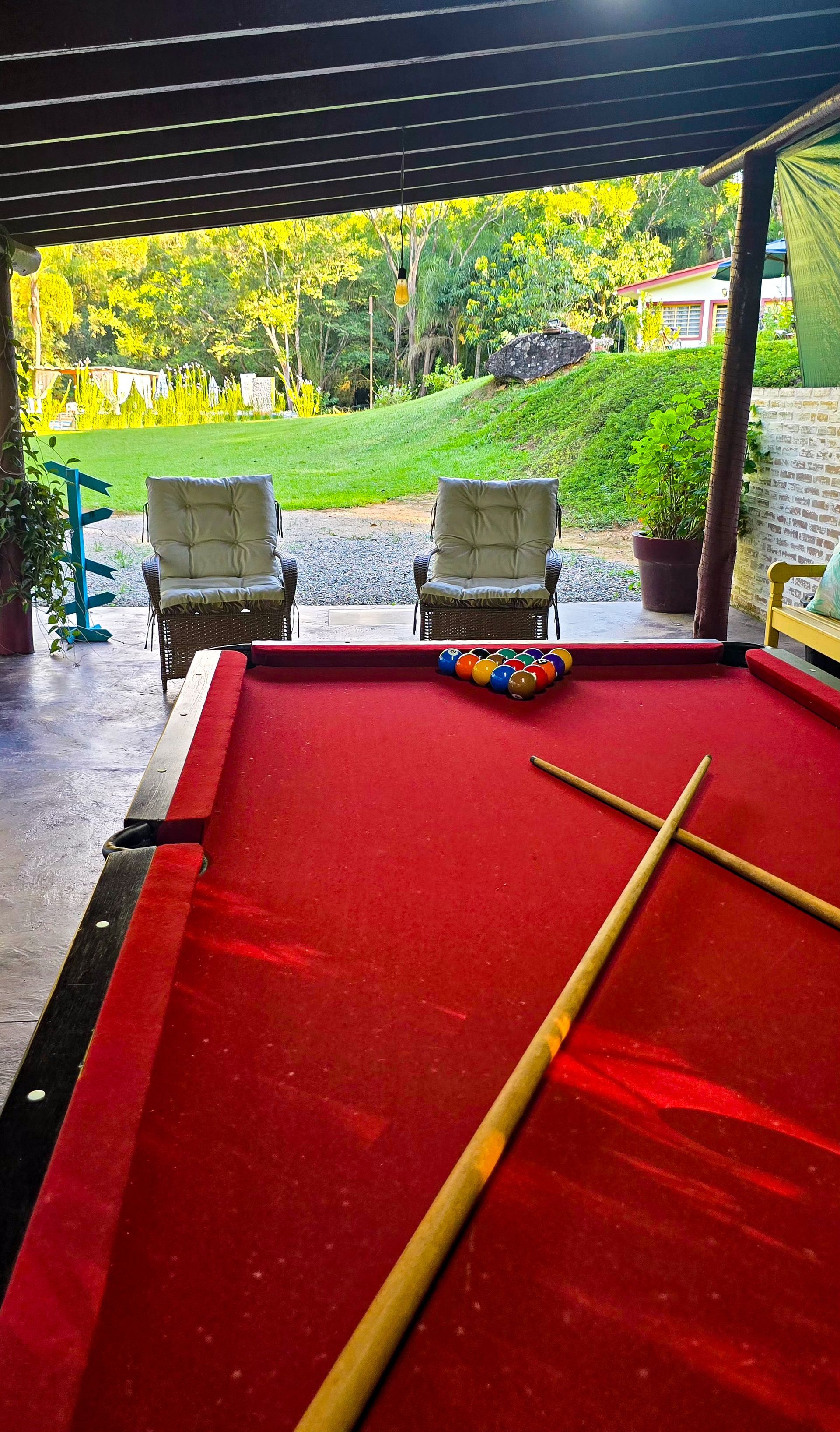 Mesa de bilhar vermelha com bolas e tacos, duas cadeiras voltadas para uma área externa verde.
