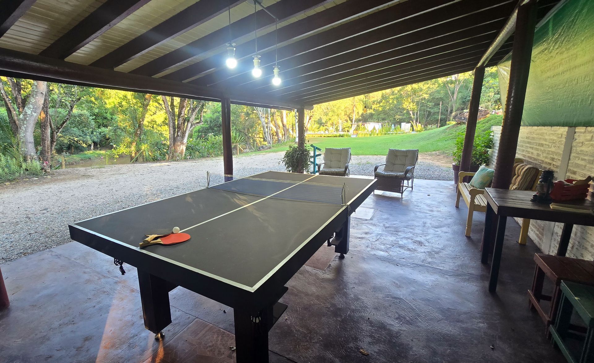 Uma mesa de pingue-pongue em um espaço externo coberto, com cadeiras, cascalho e árvores ao fundo.