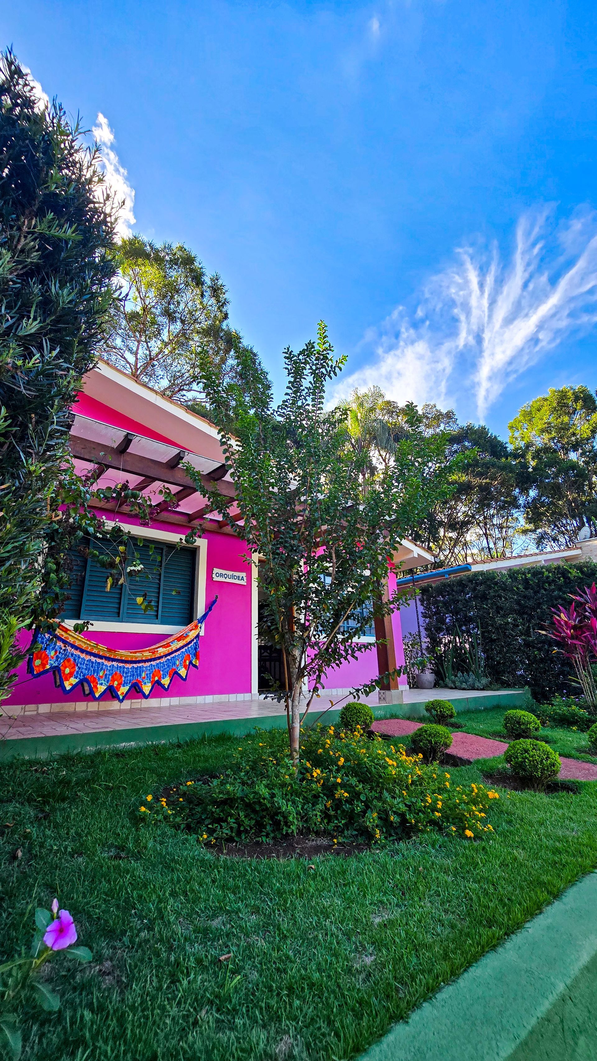Prédio rosa com gramado verde, árvores, céu azul com nuvens esparsas.