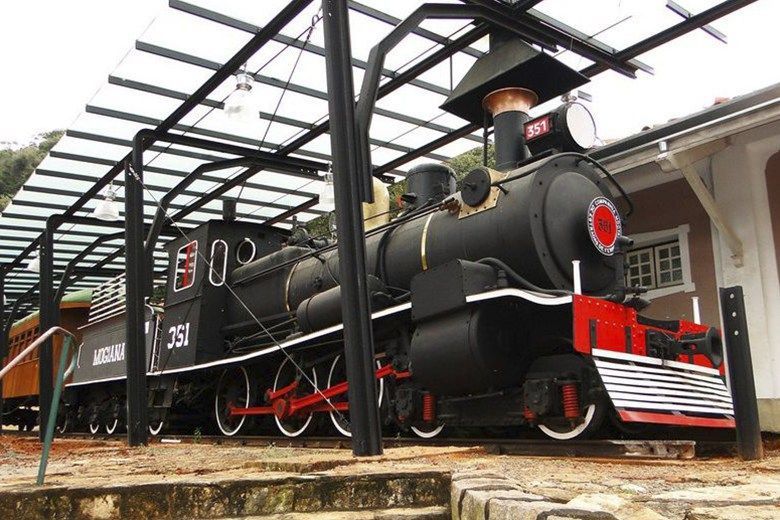 Trem a vapor preto com detalhes em vermelho e branco, sob uma plataforma coberta em uma estação.