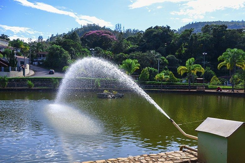 Uma fonte jorra água em um lago. Árvores verdejantes e edifícios compõem o cenário sob um céu azul.