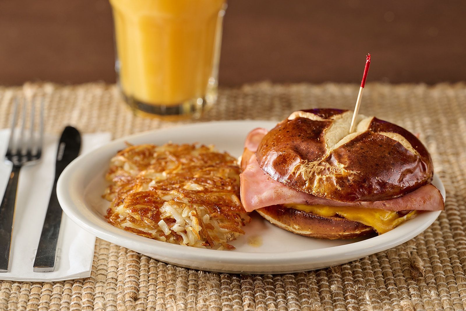 A breakfast plate with a ham and cheese pretzel bun sandwich and a side of golden-brown hash browns, plus orange juice.