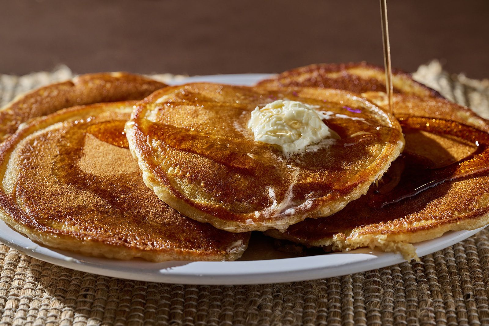 A stack of golden-brown pancakes on a white plate, topped with melting butter as syrup is drizzled over them.