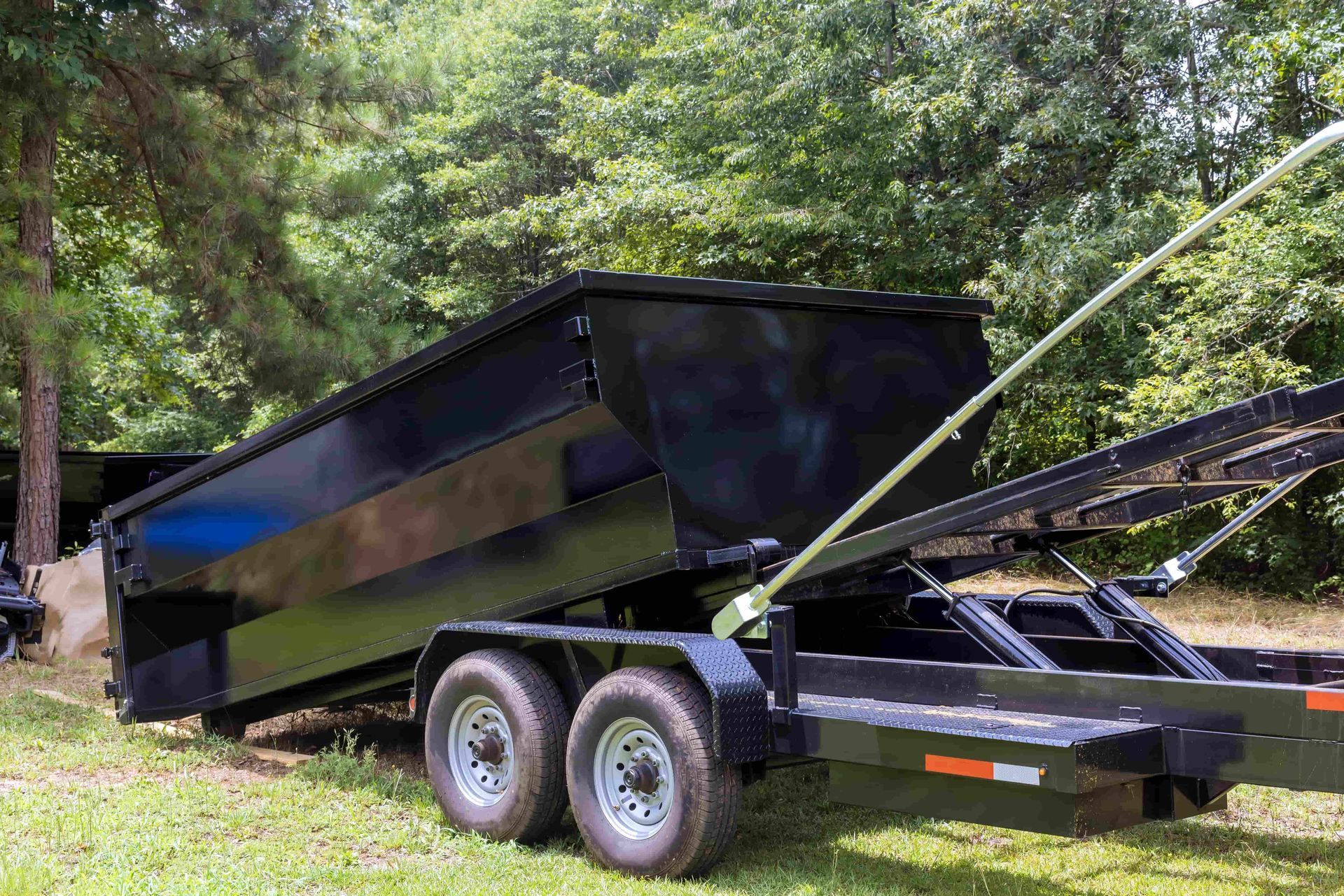 Black dump trailer parked on grass with its bed raised in a wooded area