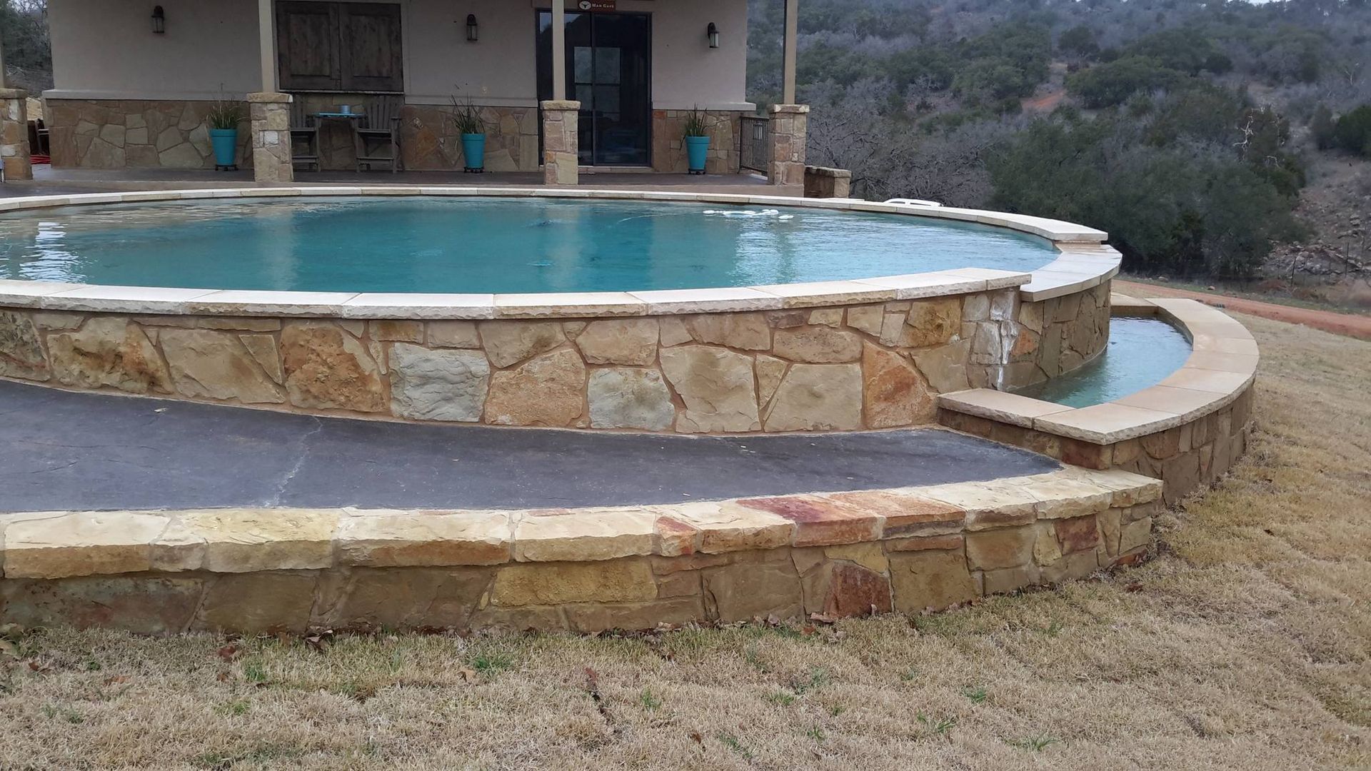 A large swimming pool is surrounded by a stone wall in front of a house.