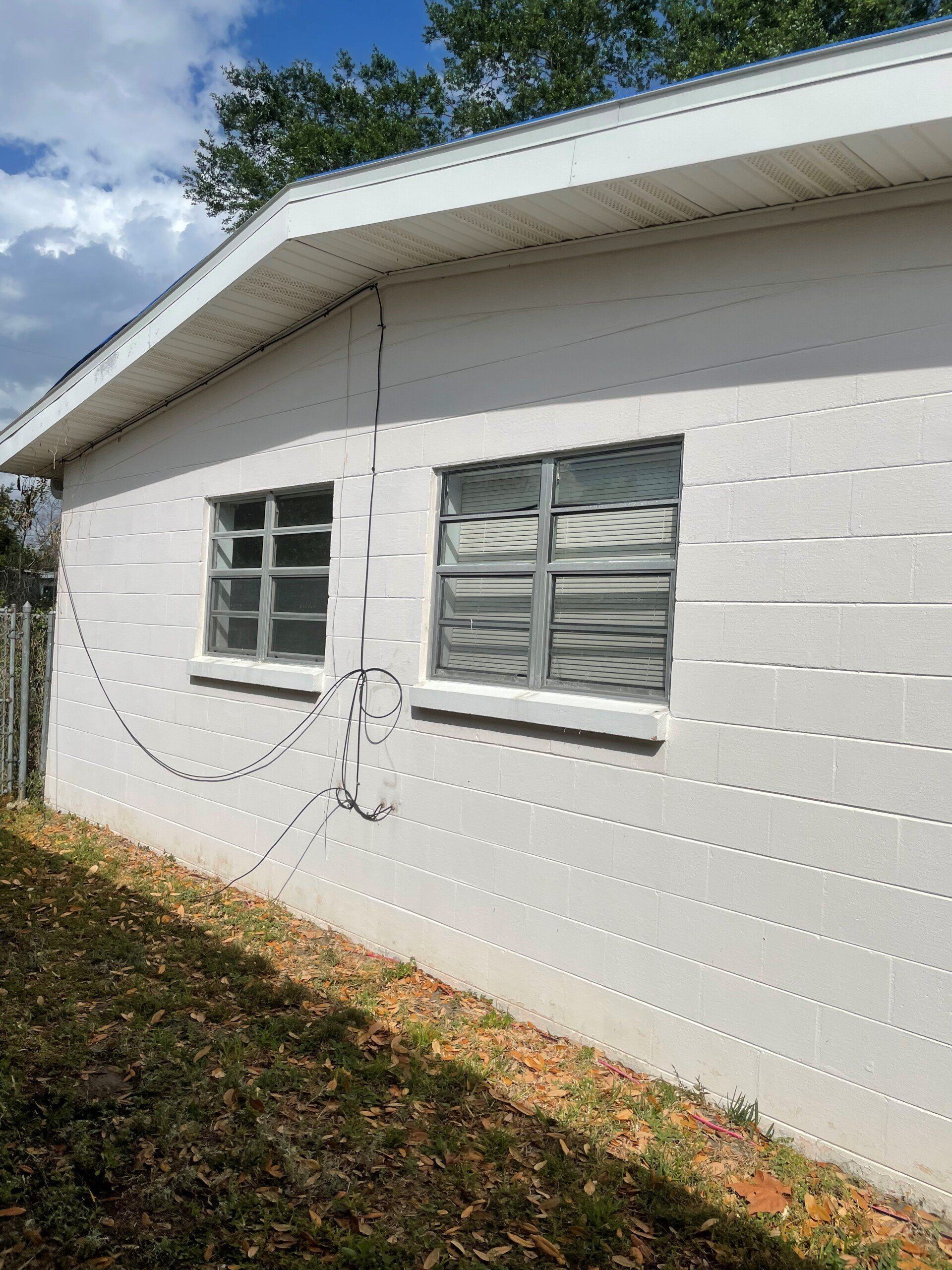 Exterior side view of a white, concrete block house with two gray-framed jalousie windows and dangling electrical wires.