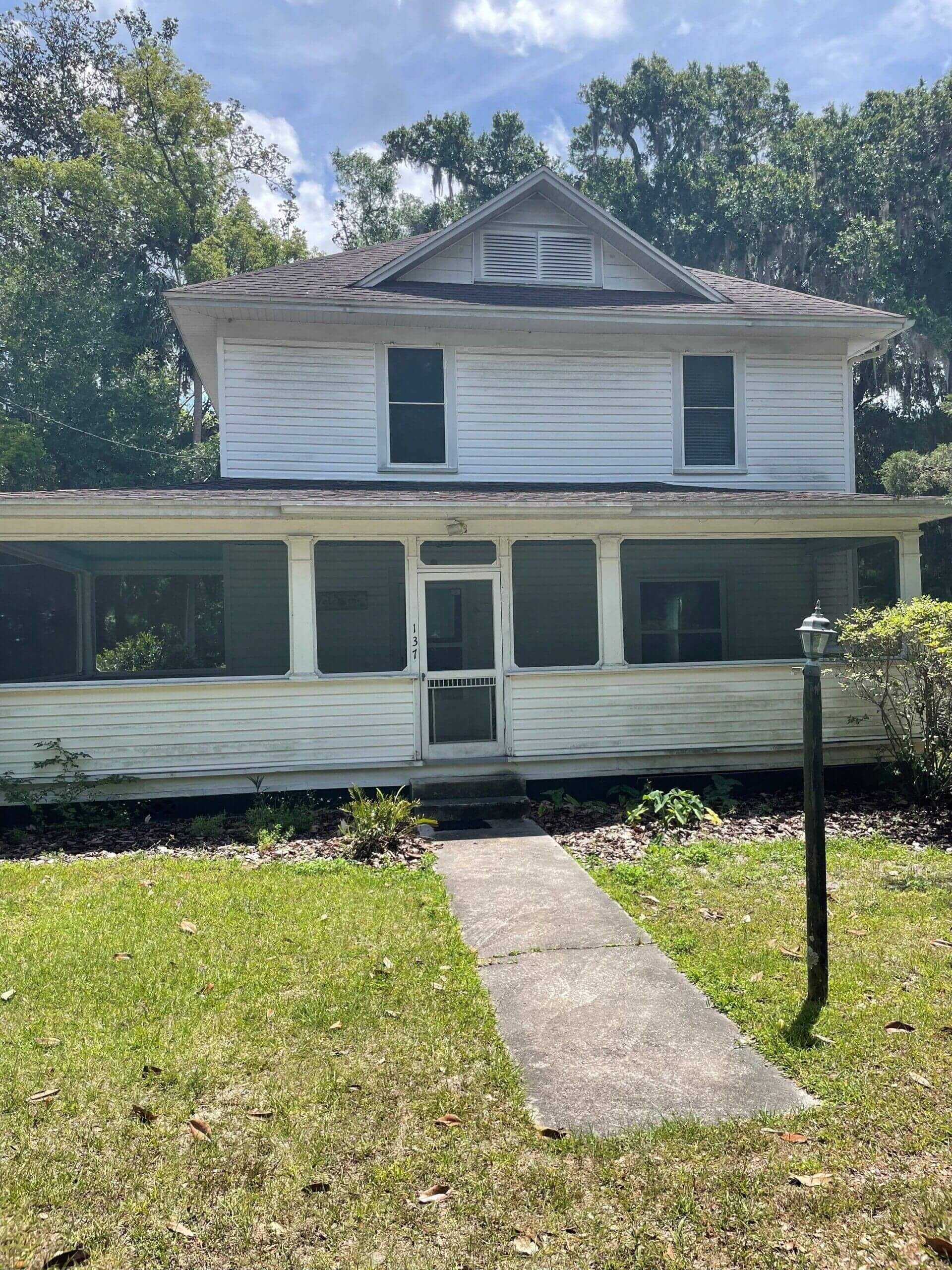 A two-story white house with a wrap-around porch and a central front door, set back from a walkway and a grassy lawn.