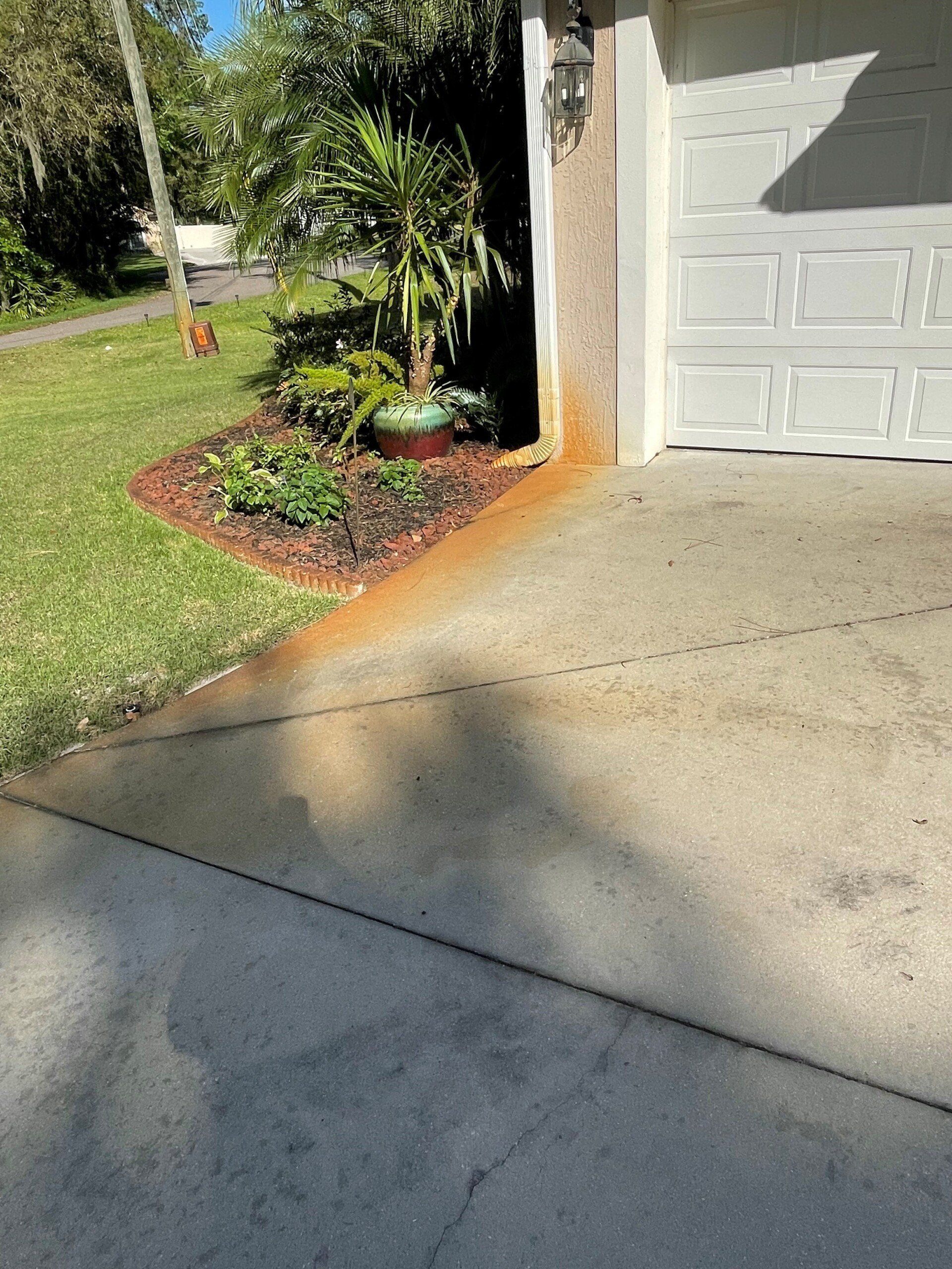 Orange rust stain from a downspout runoff on a concrete driveway near a house exterior wall and a landscaped garden bed.