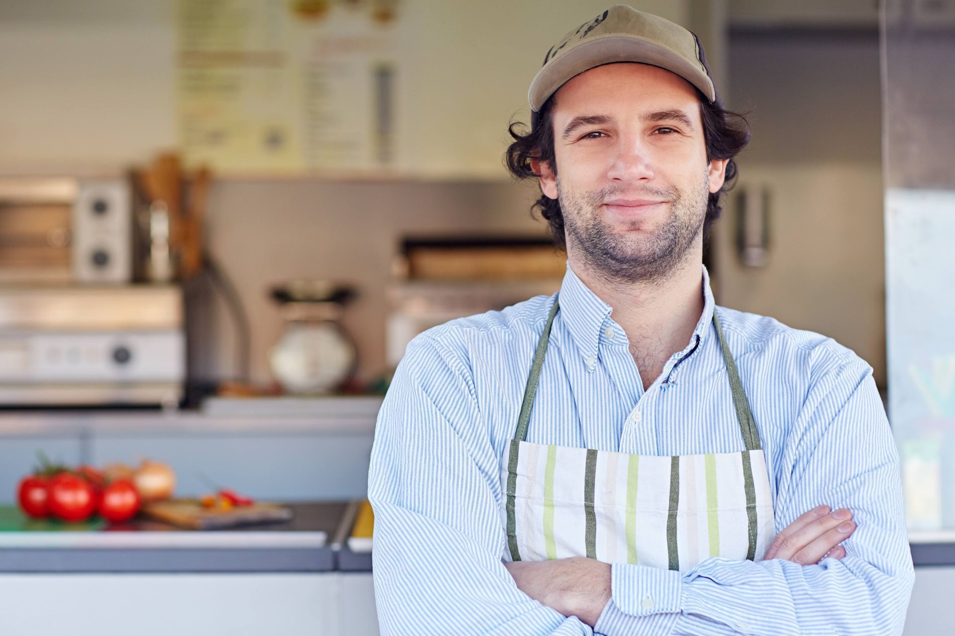 Man with arms crossed, wearing apron and cap, in front of food truck with tomatoes and onions.