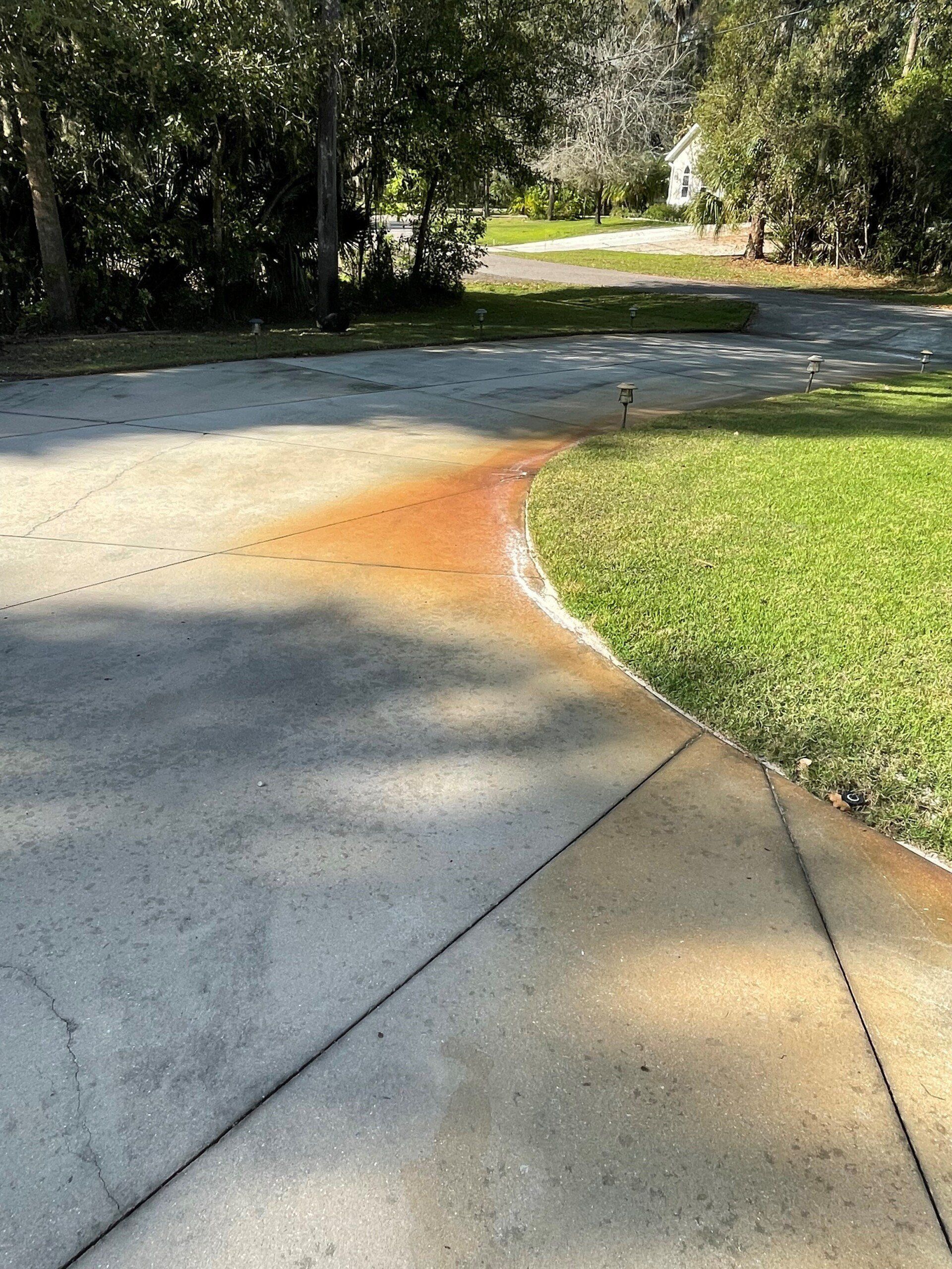 A concrete driveway with a large, bright orange rust stain running along the edge of the adjacent green lawn.