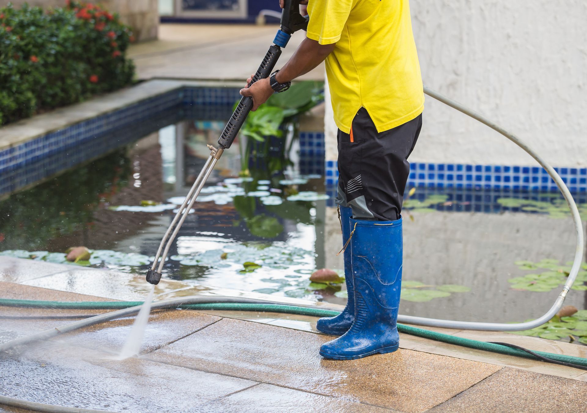 A person in a yellow shirt and blue boots uses a pressure washer to clean a tiled patio near a pond.