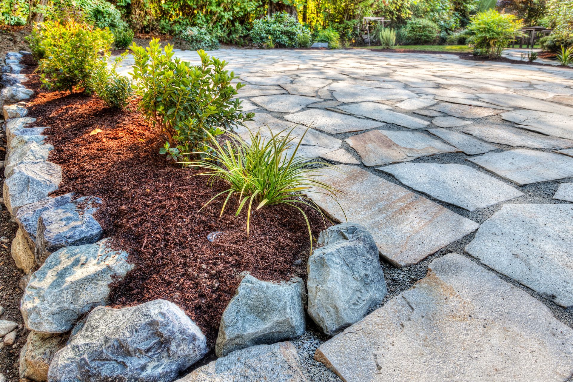 A garden bed with mulch and small green plants edged by large grey rocks next to a stone patio.