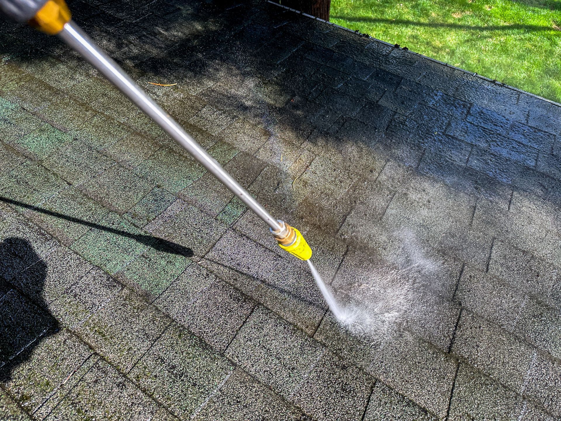 A pressure washer wand sprays a stream of water onto a stained, weathered shingled roof, cleaning a dark patch.