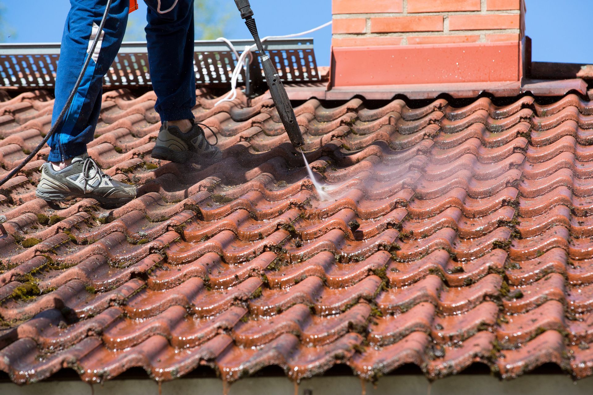 A person power-washing moss and debris off a red tiled roof near a chimney.