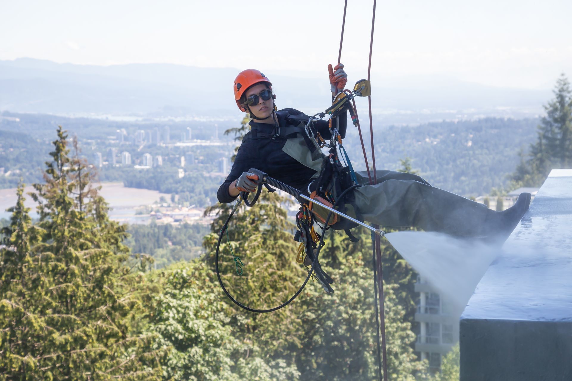 A rope access worker is soft-washing a commercial roof while suspended by a harness and safety lines.