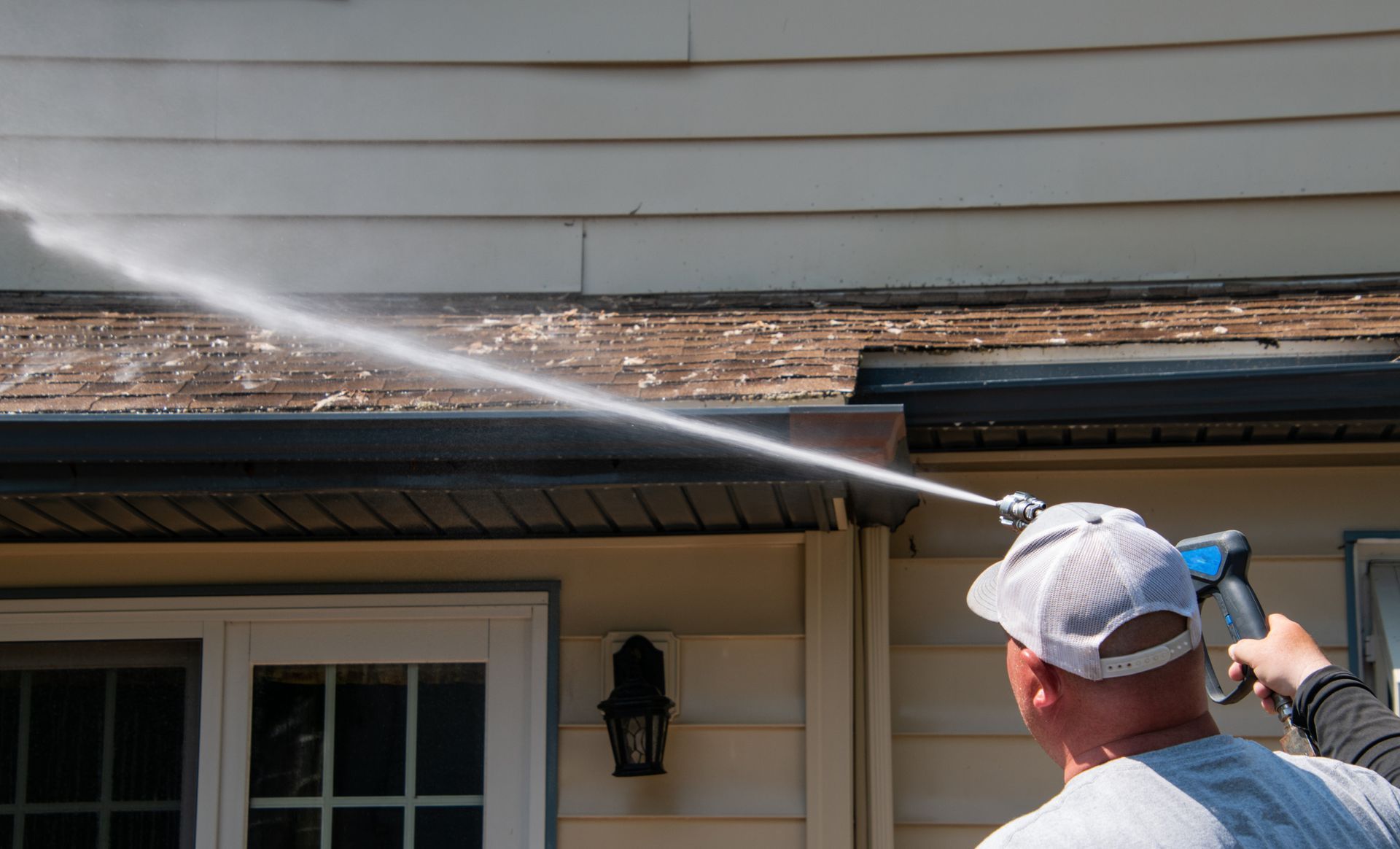 A person wearing a baseball cap uses a pressure washer to clean the exterior of a light-colored house.