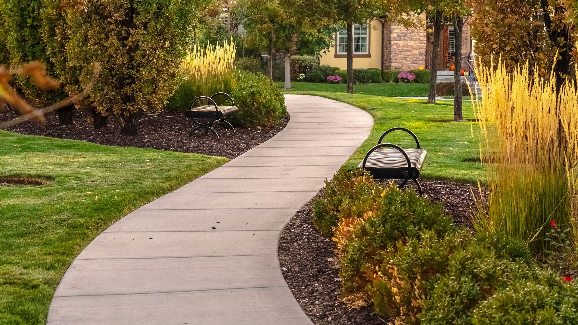 A curved concrete pathway winds through a park with green grass, landscaping, and metal benches.